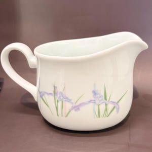 May include: A white ceramic creamer pitcher featuring a delicate floral pattern of light purple iris flowers with green stems and faint vertical pastel stripes. The pitcher has a curved handle and a wide pouring spout, with a smooth finish.
