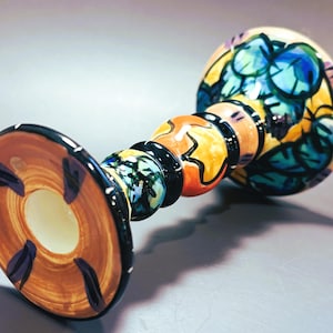 May include: A ceramic candlestick holder featuring a flared base with brown and black accents. The stem consists of stacked, rounded segments in orange, black, and blue-green floral patterns, leading to a wide, bowl-shaped top with blue leaves.