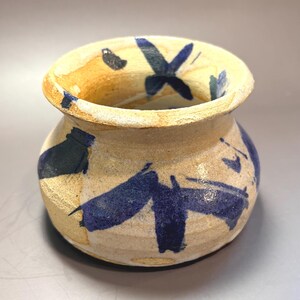 May include: A small, round ceramic pot with a wide, flared rim. The vessel features a textured, light beige clay body decorated with bold, hand-painted dark blue brushstrokes in abstract patterns across the exterior and inner rim area.