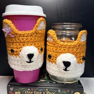 May include: Two crochet cup sleeves shaped like fox faces in orange and white yarn. The left sleeve is on a pink cup with a white lid and a small purple bow. The right sleeve is on a clear glass jar. Both feature black eyes and noses.