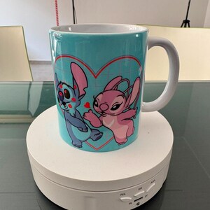 May include: A light blue ceramic mug featuring a graphic print of two cartoon characters, one blue and one pink, positioned inside a red heart outline. The mug has a white interior and a white handle, showcasing a grid pattern background design.
