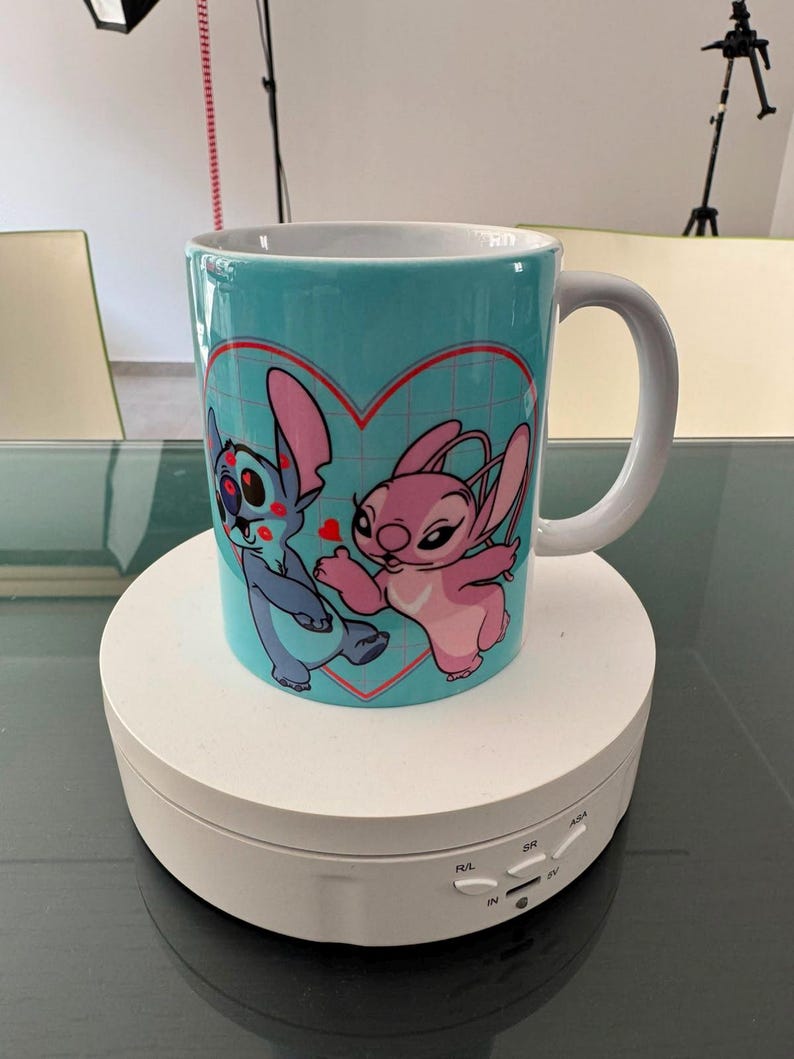May include: A light blue ceramic mug featuring a graphic print of two cartoon characters, one blue and one pink, positioned inside a red heart outline. The mug has a white interior and a white handle, showcasing a grid pattern background design.