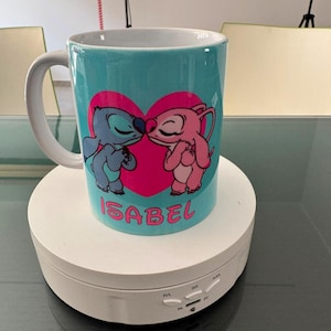 May include: A light blue ceramic mug featuring a graphic of two cartoon characters kissing in front of a large pink heart. The name ISABEL is printed in pink capital letters below the heart. The mug has a white handle and a white interior.