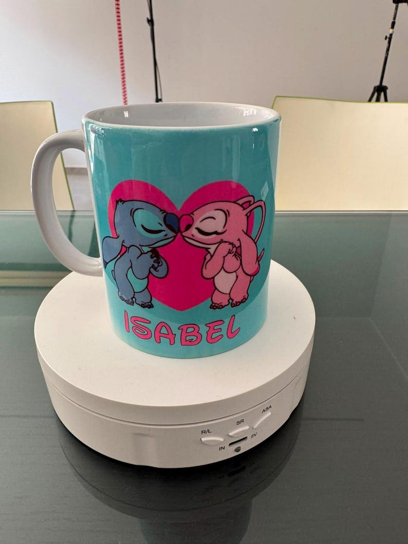 May include: A light blue ceramic mug featuring a graphic of two cartoon characters kissing in front of a large pink heart. The name ISABEL is printed in pink capital letters below the heart. The mug has a white handle and a white interior.