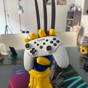 May include: A white Battletron video game controller held by a yellow and blue hand-shaped stand with three long, dark gray claws extending upward from the fingers. The controller features black analog sticks, a directional pad, and lettered buttons.