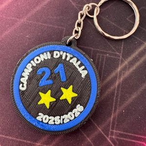May include: A circular keychain featuring a black center with the blue number 21 and two yellow stars. The border is blue with the white text CAMPIONI D'ITALIA and 2025/2026. The keychain includes a silver metal split ring attached by a short chain.