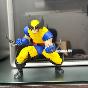 Può includere: Un action figure di Wolverine in posa da combattimento accovacciata, che indossa una tuta gialla e blu con una cintura rossa. Il personaggio presenta una maschera nera con orecchie a punta, stivali neri e artigli color argento su entrambe le mani.