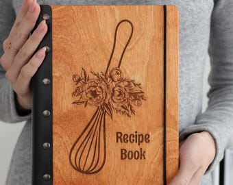 May include: A rectangular recipe book with a light brown wooden cover featuring a laser-engraved whisk adorned with floral details and the text Recipe Book. The spine is black with five silver metal rivets and a black elastic band closure.