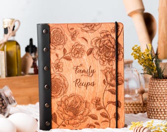 Peut inclure: Un livre de recettes en bois avec une finition brun clair, orné de motifs floraux gravés au laser et du texte "Family Recipes" en écriture cursive. La tranche est noire avec cinq rivets métalliques et un élastique noir.