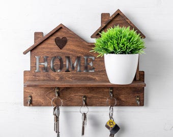 Peut inclure: Un porte-clés mural en bois brun foncé en forme de maison avec un cœur découpé et le mot HOME en lettres métalliques. Il comprend une petite étagère supportant un pot blanc avec une plante artificielle verte et cinq crochets métalliques.