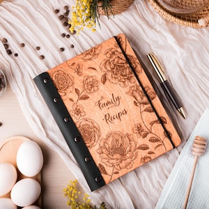 May include: A wooden recipe book with a light brown finish, featuring an engraved floral pattern and the text "Family Recipes" on the cover. The book has a black spine secured with five metal rivets and a black elastic band closure.