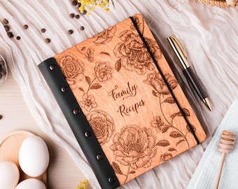 Peut inclure: Un livre de recettes en bois avec une finition brun clair, orné d'un motif floral gravé et du texte "Family Recipes" sur la couverture. Le livre possède une reliure noire fixée par cinq rivets métalliques et une fermeture élastique noire.