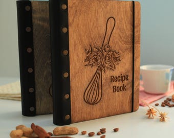 Peut inclure: Deux livres de recettes en bois avec des dos en cuir marron foncé fixés par des rivets métalliques. La couverture présente une gravure laser d'un fouet orné d'un bouquet floral et le texte Recipe Book. Un élastique noir maintient la couverture.