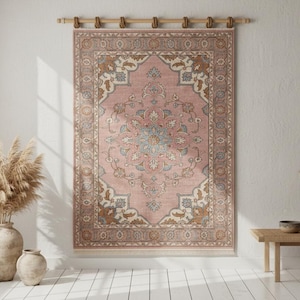 May include: A rectangular wall hanging rug featuring a dusty pink background with intricate floral and vine patterns in shades of blue, cream, and brown. The rug is suspended from a wooden rod with fabric loops along the top edge and has fringe.