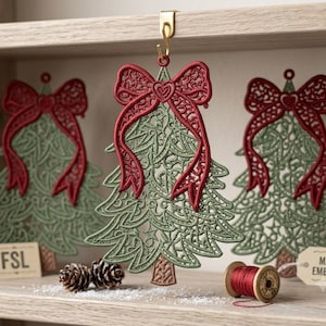 K&ouml;nnte beinhalten: Drei freistehende Weihnachtsbaumschmuck-Anh&auml;nger aus Spitze mit filigraner salbeigr&uuml;ner Stickerei, einer auff&auml;lligen roten Schleife mit Herzdetail an der Spitze und einem braunen Stamm. Ein Anh&auml;nger h&auml;ngt an einem goldenen Haken, zwei weitere stehen daneben mit Schildern, die FSL und MACHINE EMBROIDERY beschriftet sind.