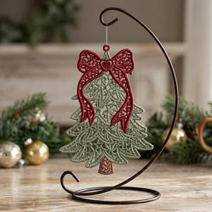 K&ouml;nnte beinhalten: Ein salbeigr&uuml;ner Weihnachtsbaumschmuck aus Spitze mit einer dunkelroten, bestickten Schleife an der Spitze und einem kleinen Herzdetail. Der Anh&auml;nger h&auml;ngt an einem d&uuml;nnen, durchsichtigen Faden an einem geschwungenen, dunkelbraunen Metallst&auml;nder.