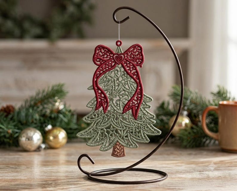 K&ouml;nnte beinhalten: Ein salbeigr&uuml;ner Weihnachtsbaumschmuck aus Spitze mit einer dunkelroten, bestickten Schleife an der Spitze und einem kleinen Herzdetail. Der Anh&auml;nger h&auml;ngt an einem d&uuml;nnen, durchsichtigen Faden an einem geschwungenen, dunkelbraunen Metallst&auml;nder.