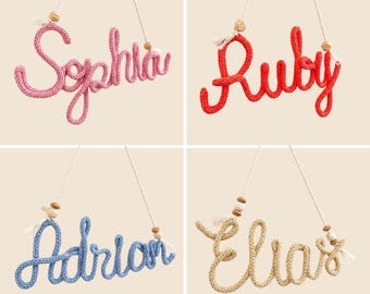 Könnte beinhalten: Vier hängende Wandschilder aus gestricktem Garn in Schreibschrift, die die Namen Sophia in Pink, Ruby in Rot, Adrian in Blau und Elias in Beige bilden. Jedes Schild hat eine weiße Schnur mit kleinen Holzperlen und Quasten.