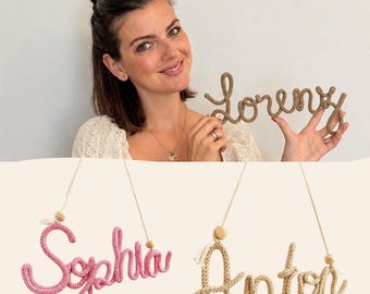 Könnte beinhalten: Personalisierte Namensschilder aus gestricktem Seil in Schreibschrift. Ein Schild zeigt den Namen Sophia in Rosa, ein weiteres Anton in Beige und ein drittes Lorenz in Braun, jeweils an einer Schnur mit einer Holzperle aufgehängt.