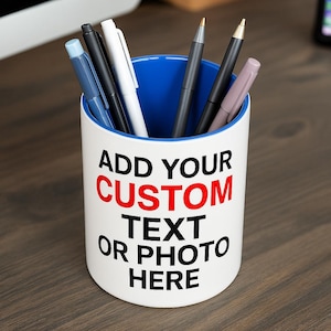 May include: A white cylindrical pen holder with a vibrant blue interior. The exterior features the black and red printed text: ADD YOUR CUSTOM TEXT OR PHOTO HERE. Several pens in various colours, including black, white, blue, and grey, are stored inside.