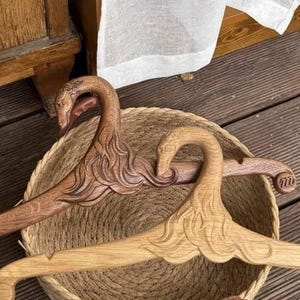 Pode incluir: Dois cabides de madeira com designs esculpidos de pescoço de cisne. Um cabide é feito de madeira castanha escura, enquanto o outro tem um tom de madeira natural mais claro. Ambos os cabides têm braços curvos e lisos dentro de uma cesta redonda.