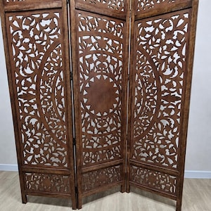 Puede incluir: Un biombo plegable de tres paneles hecho de madera marrón con intrincados patrones florales y geométricos cortados con láser. Cada panel tiene un borde superior festoneado y un motivo circular central, unidos por bisagras de metal negro.