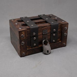 May include: A rectangular dark brown wooden chest featuring two vertical matte black metal straps secured with rounded rivets. The lid is fastened with a matching metal latch and a small, silver-toned padlock hanging from the front center.