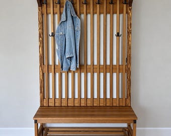 May include: A tall wooden hall tree featuring a slatted back panel with multiple black metal hooks, a flat bench seat, and two lower slatted shelves for shoe storage. A light blue denim jacket hangs from one of the upper hooks on the unit.