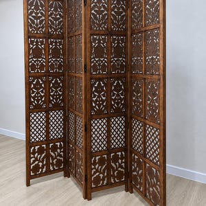 May include: A three-panel folding room divider made of dark brown wood. Each panel features a rectangular frame divided into sections with intricate laser-cut floral and geometric lattice patterns. The panels are connected by black metal hinges.