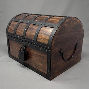 May include: A dark brown wooden chest with a curved lid, featuring black metal bands and decorative rivets. The chest includes a black metal handle on the side and a black padlock secured to the front latch, providing a sturdy storage container.