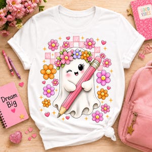 Peut inclure: Un t-shirt blanc avec un motif représentant un fantôme qui fait un clin d'œil, portant une couronne de fleurs colorées et tenant un grand crayon rose. Le design est entouré de fleurs roses et orange, de petits cœurs, d'étoiles jaunes et d'un motif à carreaux rose.