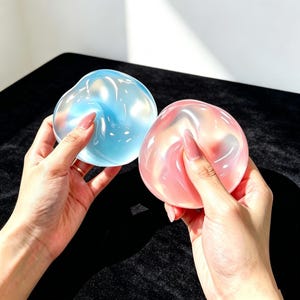 May include: Two circular, translucent silicone objects with a smooth, pliable texture. One object is a light, icy blue colour, and the other is a soft, pale pink. Both items have a glossy, reflective surface that catches the light.