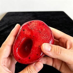 K&ouml;nnte beinhalten: Ein glattes, gl&auml;nzendes, leuchtend rotes Harzobjekt in Form einer halbierten Frucht mit einer tiefen zentralen Vertiefung und kleinen schwarzen Sprenkeln auf der Oberfl&auml;che, die das Aussehen von Samen in einem frischen Obstst&uuml;ck nachahmen.