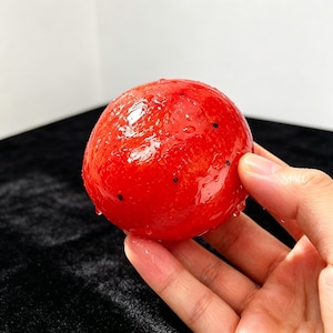 K&ouml;nnte beinhalten: Eine runde, leuchtend rote Frucht mit einer gl&auml;nzenden, nassen Oberfl&auml;chenstruktur und kleinen, verstreuten schwarzen Punkten. Die Frucht ist kugelf&ouml;rmig und wirkt frisch gewaschen, mit winzigen Wassertropfen auf ihrer lebendigen, glatten Haut.