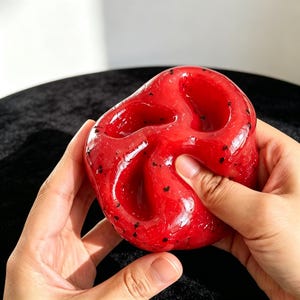 Może przedstawiać: Błyszczący, półprzezroczysty czerwony slime o gęstej, żelowej konsystencji z zatopionymi małymi czarnymi drobinkami. Materiał jest elastyczny i podatny na formowanie, z widocznymi głębokimi wgłębieniami i fałdami powstałymi przez ściskanie.