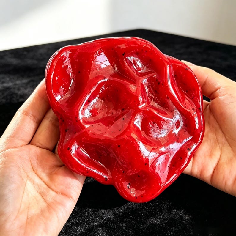 K&ouml;nnte beinhalten: Eine gl&auml;nzende, leuchtend rote Keramikschale mit einer unregelm&auml;&szlig;igen, organischen und wellenf&ouml;rmigen Form. Die Oberfl&auml;che weist ein glattes, reflektierendes Finish mit feinen dunklen Sprenkeln auf, die im Material eingebettet sind.