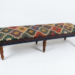 May include: A long rectangular bench featuring a woven fabric top with a repeating diamond pattern in shades of red, green, blue, and yellow. The bench is supported by six turned wooden legs with a dark brown finish and a dark blue fabric trim.