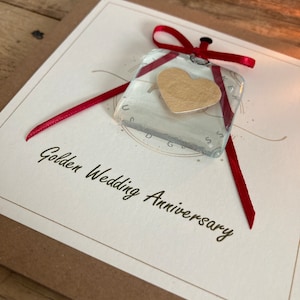 May include: A white card with the text "Golden Wedding Anniversary" and a red ribbon tied around a clear glass square with a gold heart inside.