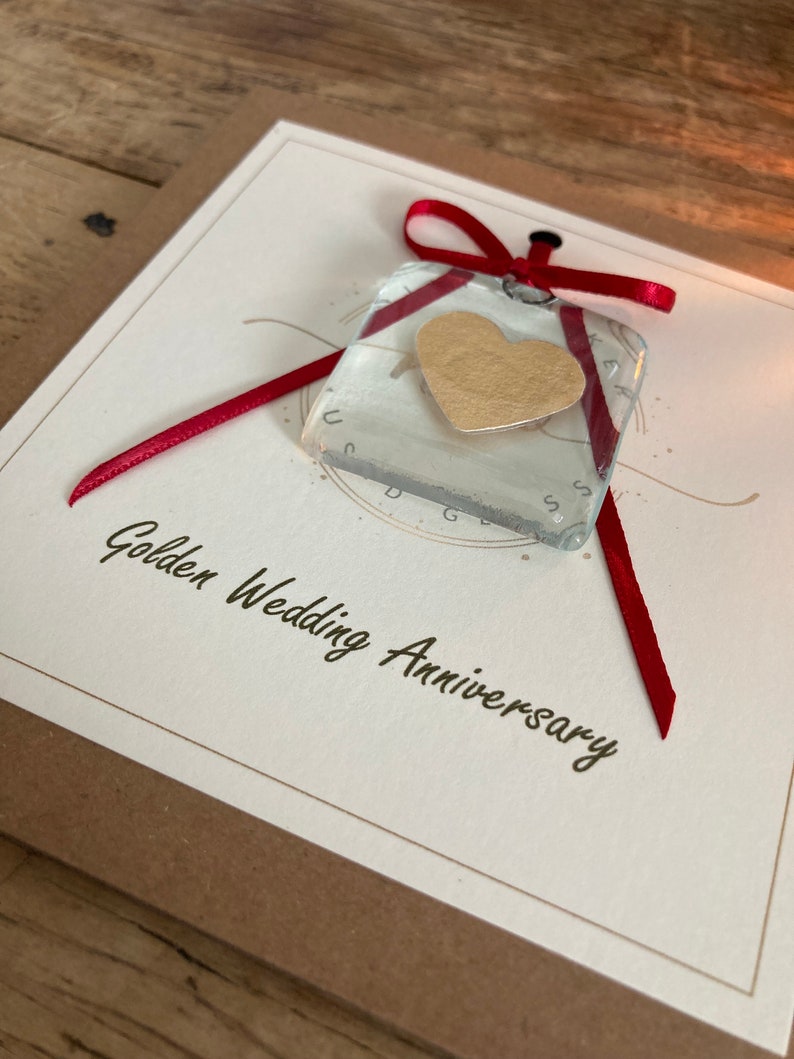 May include: A white card with the text "Golden Wedding Anniversary" and a red ribbon tied around a clear glass square with a gold heart inside.