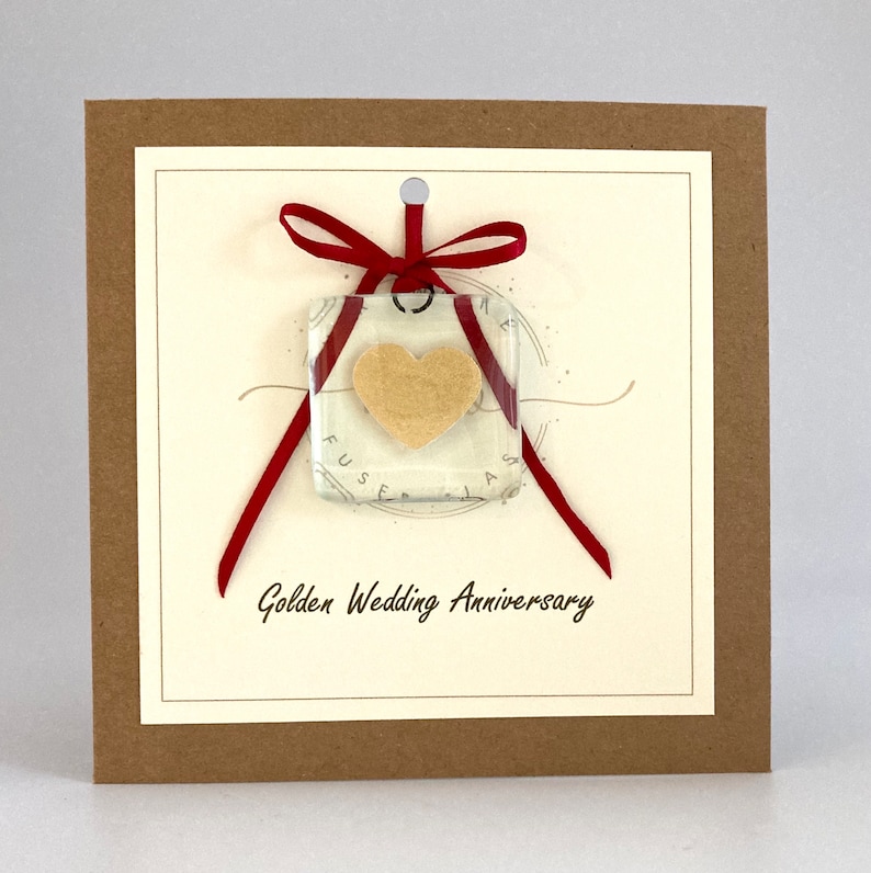 May include: A card with a red ribbon tied around a square glass ornament with a gold heart inside. The card has the text "Golden Wedding Anniversary" printed on it.