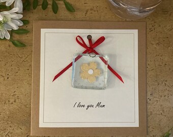 May include: A card with a red ribbon and a small glass square with a yellow flower inside. The card says "I love you Mum".