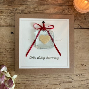May include: A card with a red ribbon tied around a clear glass square with a gold heart inside. The card has the text "Golden Wedding Anniversary" printed on it.