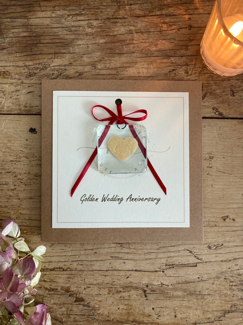 May include: A card with a red ribbon tied around a clear glass square with a gold heart inside. The card has the text "Golden Wedding Anniversary" printed on it.