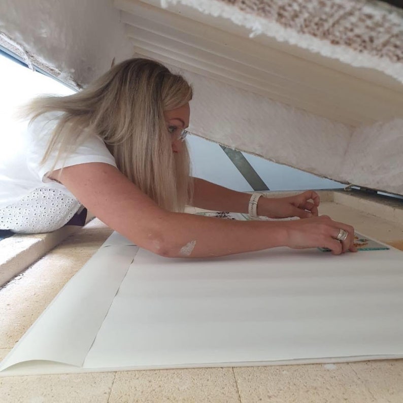 May include: A woman is working on a large white paper, possibly a map or a craft project. She is wearing a white shirt with a delicate pattern and has blonde hair.