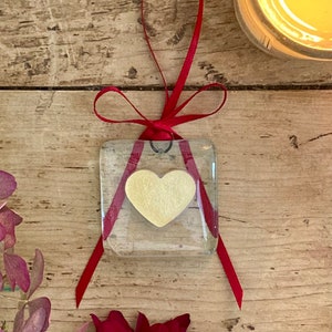 May include: A small glass square ornament with a yellow heart inside. The ornament is tied with a red ribbon and hangs from a black metal loop.