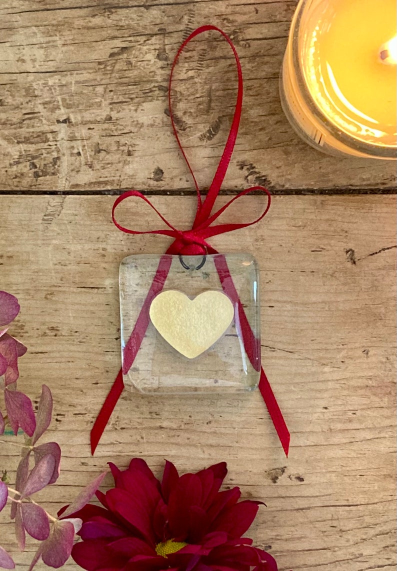 May include: A small glass square ornament with a yellow heart inside. The ornament is tied with a red ribbon and hangs from a black metal loop.