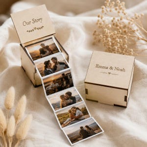 May include: A light-colored wooden box with a hinged lid engraved with the text Our Story and a decorative flourish. Inside, a connected accordion-style photo strip displays five images of couples. A second matching wooden box is engraved Emma & Noah.