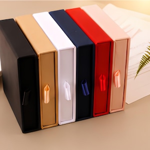 May include: A set of six rectangular drawer-style gift boxes arranged in a row. The boxes are black, gold, white, navy blue, red, and light pink. Each box features a matching satin ribbon pull tab centered on the front panel for easy opening.