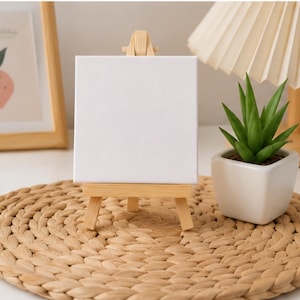 May include: A small, square, blank white canvas mounted on a light-colored wooden tripod easel. The canvas has a smooth, matte texture and is positioned upright, providing a clean surface for artistic display or creative projects.