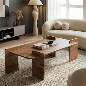 Pode incluir: Uma mesa de centro retangular de madeira com bordas curvas e pernas em formato de U, posicionada em uma sala de estar com um sofá curvo na cor creme, uma poltrona de couro marrom, um rack cinza, uma luminária de chão e um tapete texturizado.